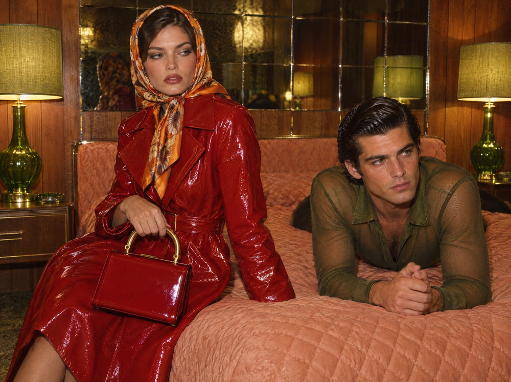 A woman in a red patent trench holding a red bag on a hotel bed, a man in a mesh shirt beside her