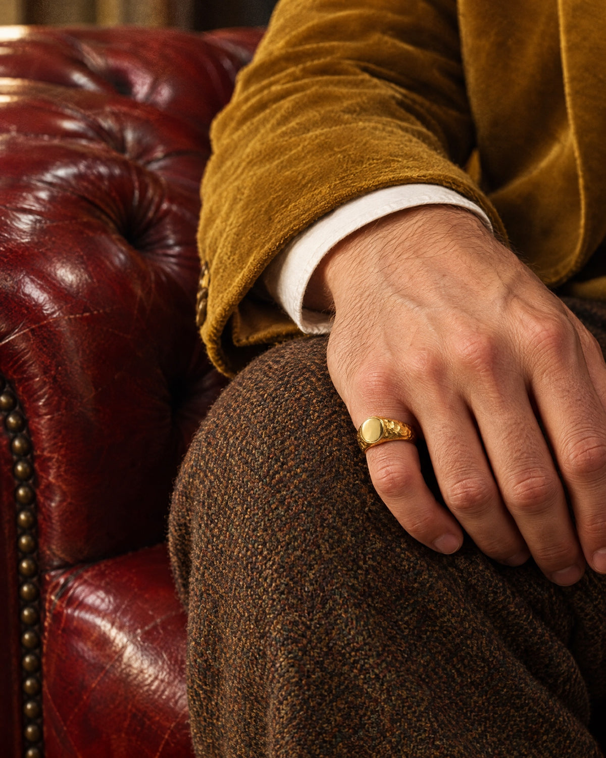 The signet ring on a man's little finger, resting on a tweed knee beside a velvet cuff